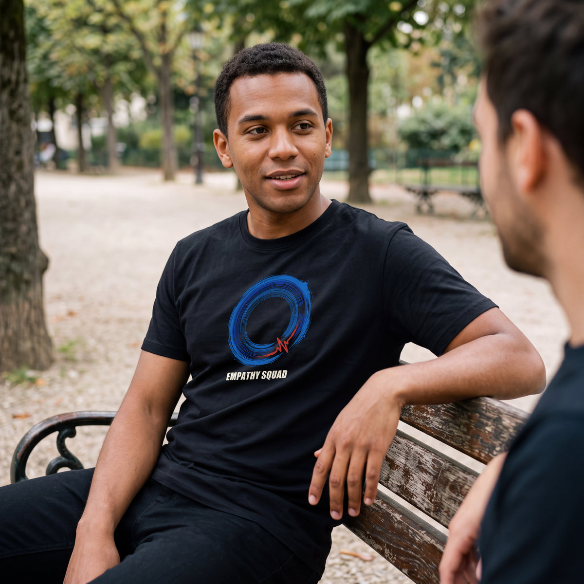 KI-generierte Produktvisualisierung: Mann im schwarzen Empathy-Squad-Shirt auf einer Parkbank im Gespräch mit einem Freund. Vollständiger blauer Kreis, roter Impuls und EMPATHY SQUAD auf der Brust.