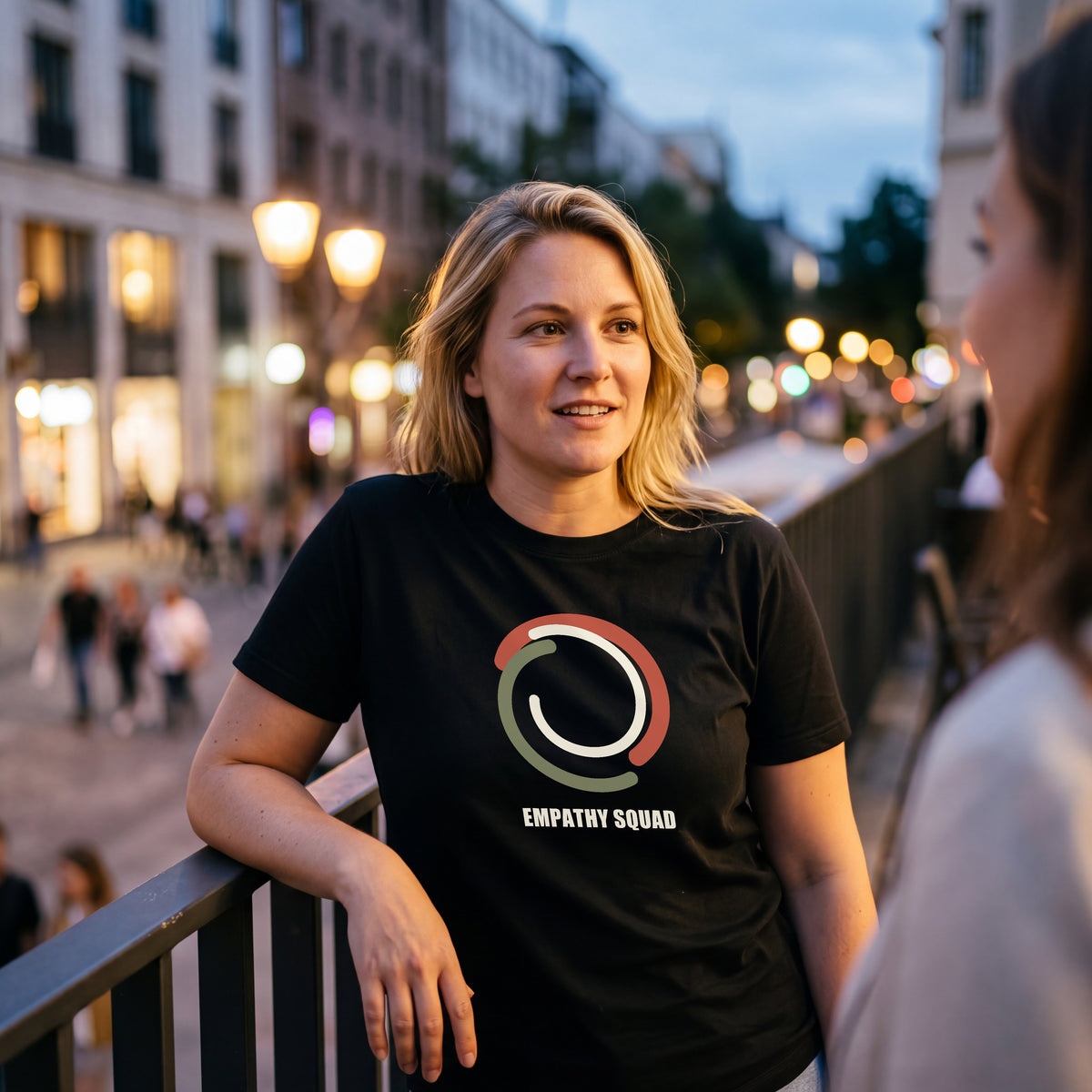 Frau beim abendlichen Treffen am Geländer im schwarzen Empathy-Squad-T-Shirt mit drei Farbbögen und vollständig sichtbarem EMPATHY SQUAD-Schriftzug auf der Brust.