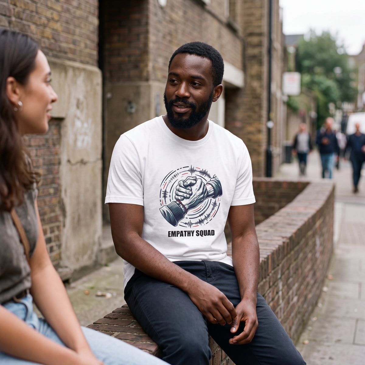 KI-erstellte Produktvisualisierung: Mann im weißen Empathy Squad Verbundenheit T-Shirt mit Händedruck-Grafik und roten Klanglinien im Gespräch vor einer Backsteinwand.