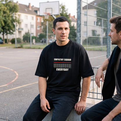 Mann am Basketballplatz im schwarzen Empathy Everyday Oversize-T-Shirt mit vollständigem EMPATHY SQUAD- und EVERYDAY-Frontprint.