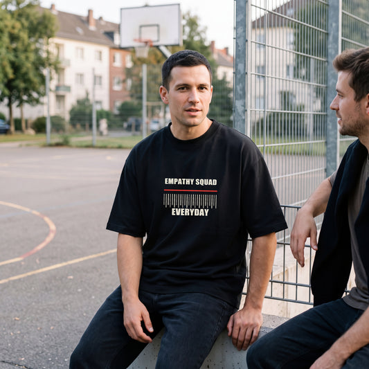 Mann am Basketballplatz im schwarzen Empathy Everyday Oversize-T-Shirt mit vollständigem EMPATHY SQUAD- und EVERYDAY-Frontprint.