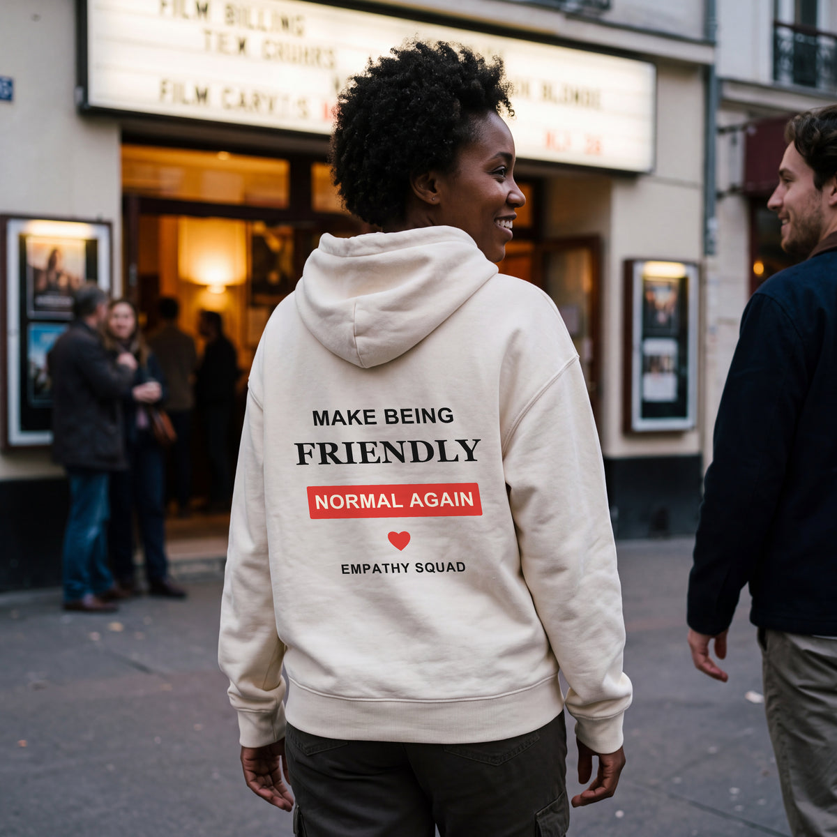 Frau vor einem Kino im Natural-Raw-Hoodie von Empathy Squad. Vollständiger Friendly-Rückenprint in Schwarz und Rot unterhalb der Kapuze; kleines Brustlogo auf dem separaten Frontbild.