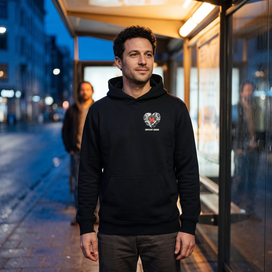 Schwarzer Empathy Squad Heart Hoodie mit kleinem Herzmotiv links auf der Brust und lesbarem EMPATHY SQUAD, getragen an einer Haltestelle am Abend.