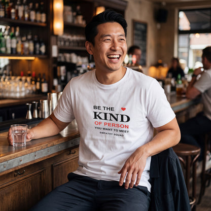 Mann in einer Bar im weißen Empathy-Squad-T-Shirt. Der vollständige schwarze und rote Schriftzug „Be the Kind of Person You Want to Meet“, das kleine Herz und EMPATHY SQUAD sitzen auf der Brust.