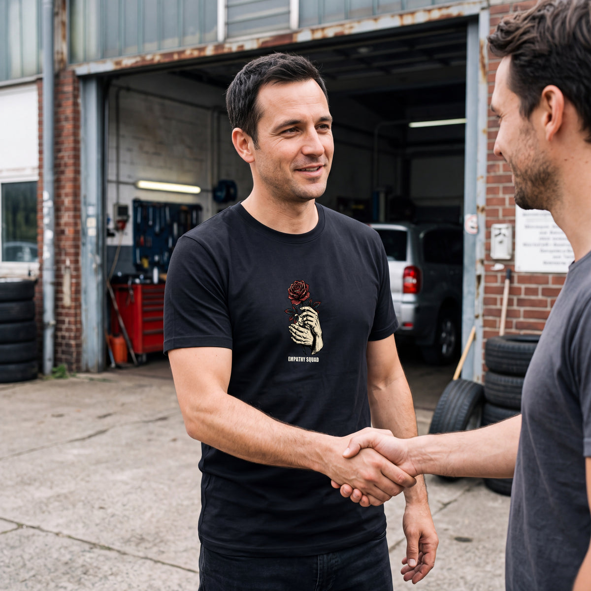 Mann vor einer Werkstatt im schwarzen Empathy-Squad-T-Shirt: zwei Hände halten eine rote Rose. Grafik und EMPATHY SQUAD-Schriftzug sind vollständig auf der oberen Brust sichtbar.